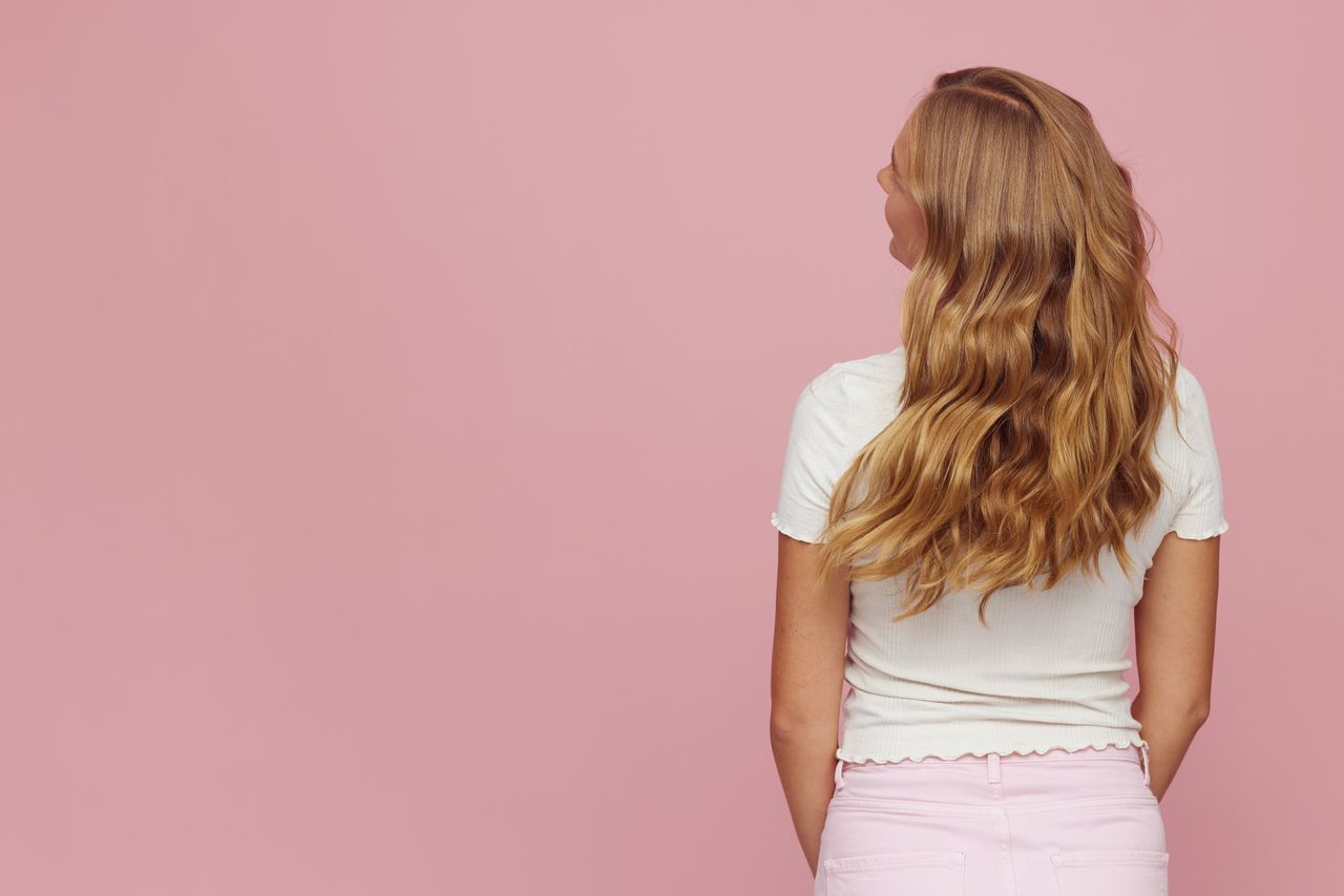 Rear view of woman with long blonde wavy hair against pink backdrop.